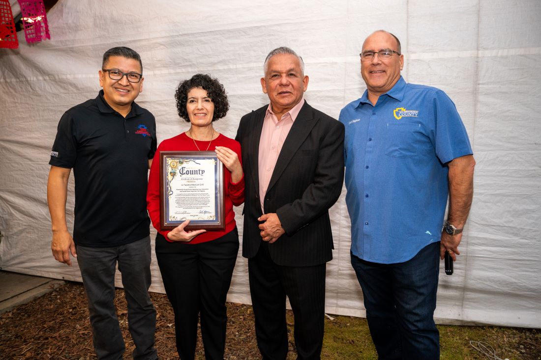 Four people pose together as a woman holds a framed County certificate at the Let's Taco 'Bout It Upland community event.