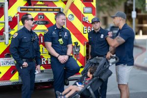 Three Chino Valley firefighters speak with a man pushing a stroller in front of a fire engine.