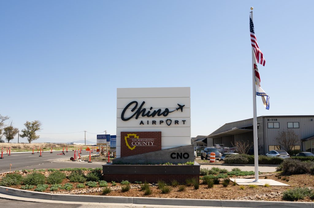 Photo of directional sign for Chino Airport. The sign is next to a flag pole with the American and California flag. The sign includes the name of the airport and the San Bernardino County Logo.