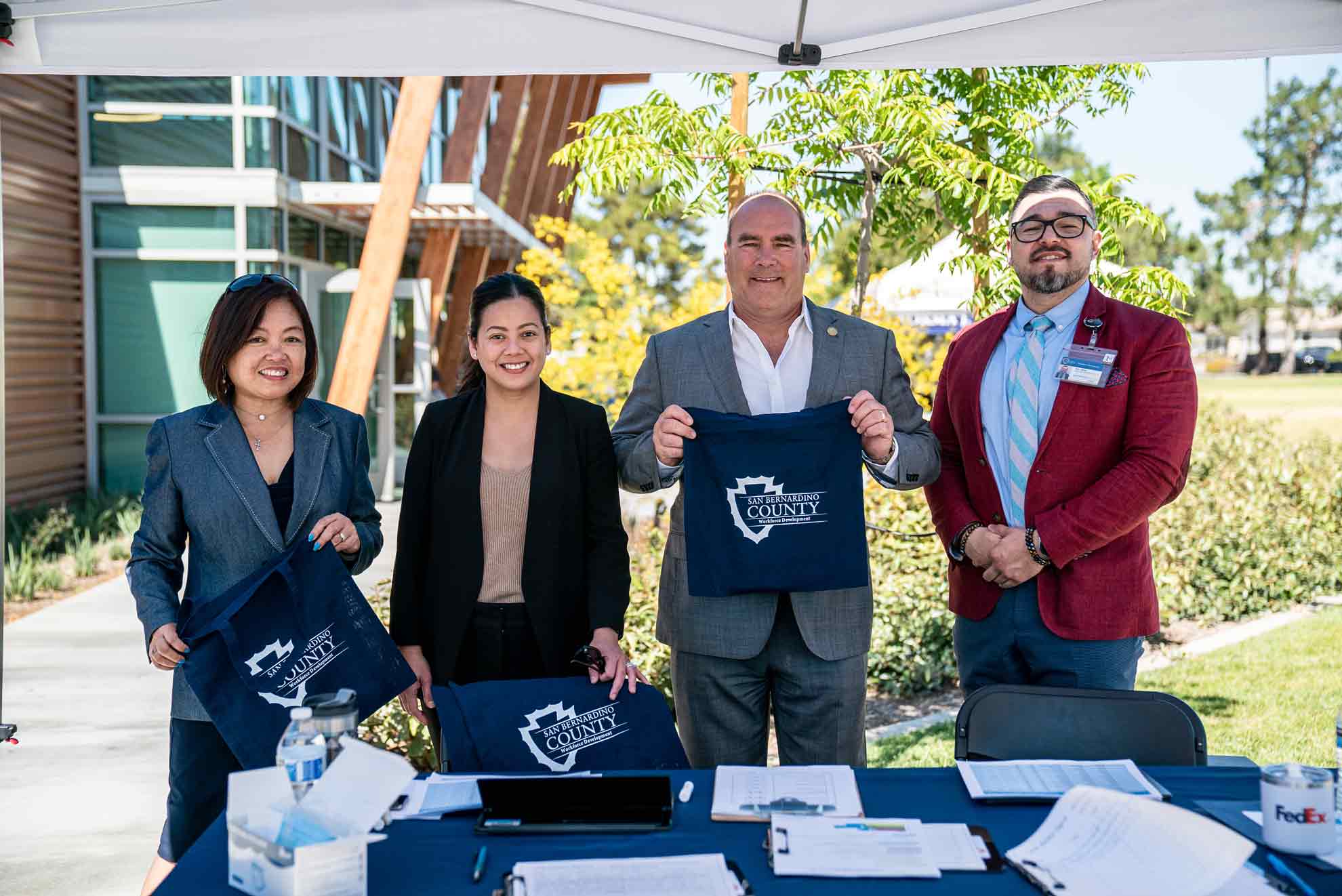 Two females and a male pose outdoors with Supervisor Hagman holding San Bernardino County tote bags at the 2022 West End Job Fair.