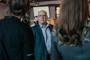 Supervisor Hagman, in a suit, speaks with others at an indoor event, with a mobile order screen visible in the background.