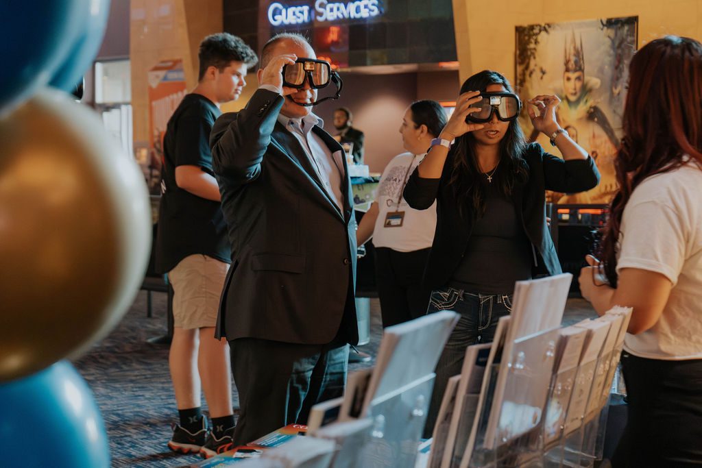 A man and a woman are trying on impairment goggles at an awareness event inside a theater lobby.