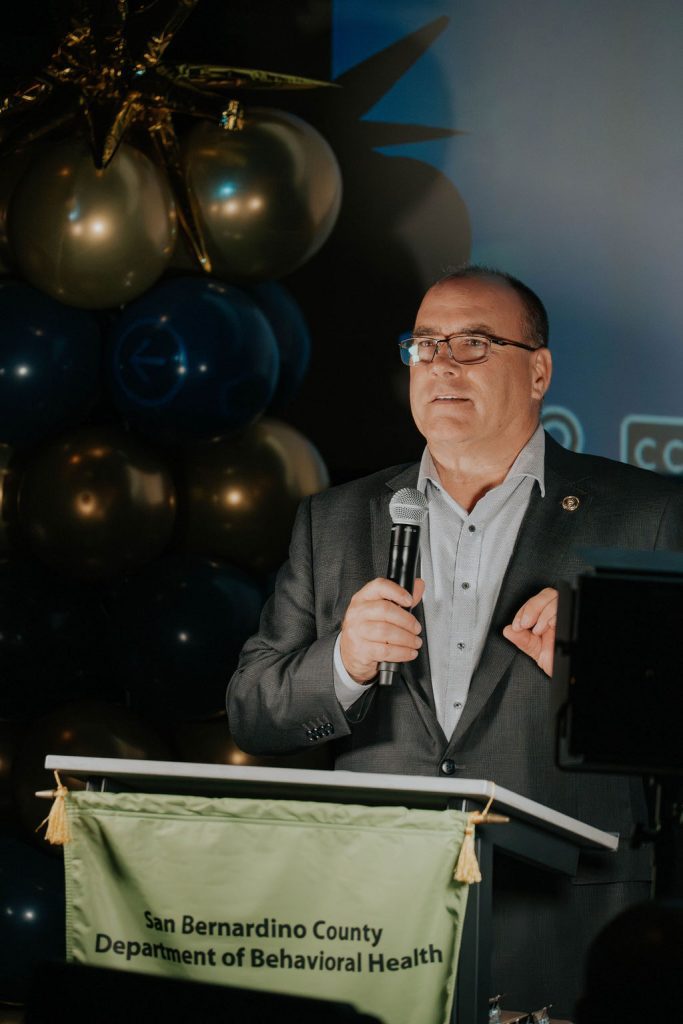 A man speaks at a podium with a San Bernardino County Department of Behavioral Health banner.