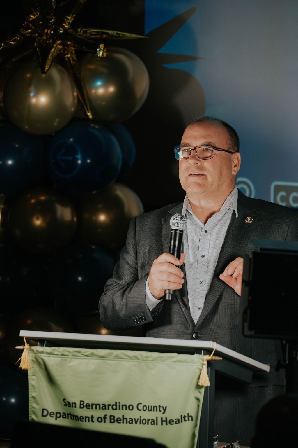 A man speaks at a podium with a San Bernardino County Department of Behavioral Health banner.