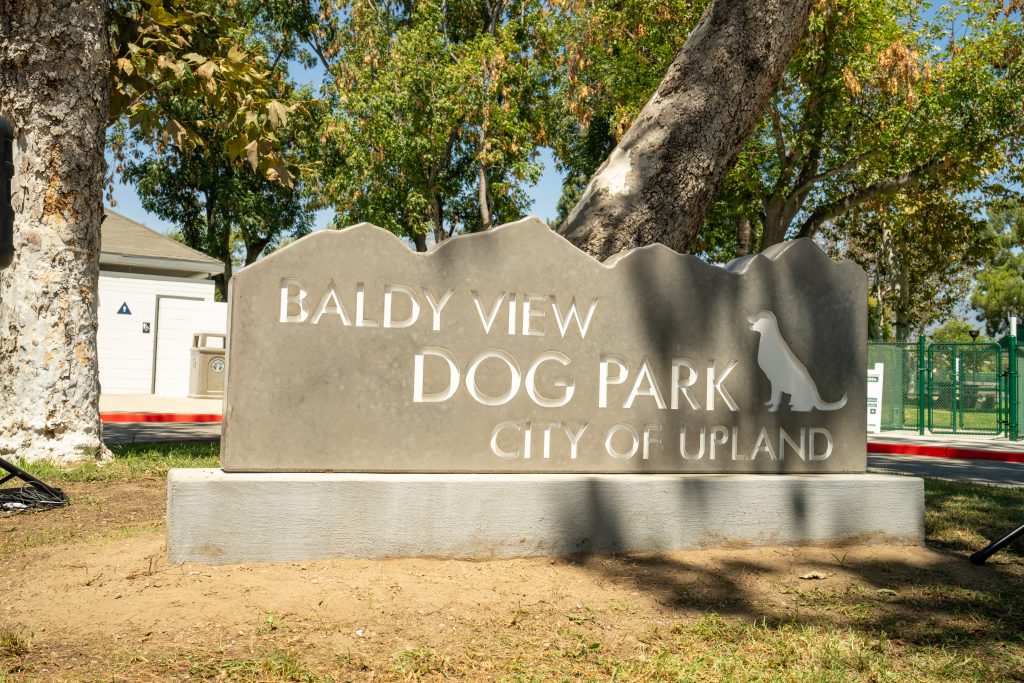 Photo of the City of Upland Baldy View Dog Park. The sign is rectangular with bumps like mountains along the top edge. The sign includes the name of the park and city along with a outline of a dog.