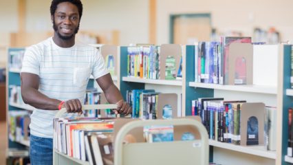 A man stands in a library, smiling while pushing a cart filled with books between bookshelves.