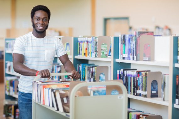 A man stands in a library, smiling while pushing a cart filled with books between bookshelves.