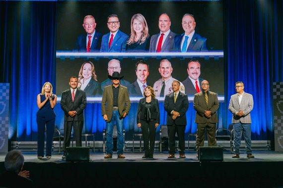 San Bernardino County Leadership stand on stage at an event, with a large screen behind them displaying portraits of community and government leaders.